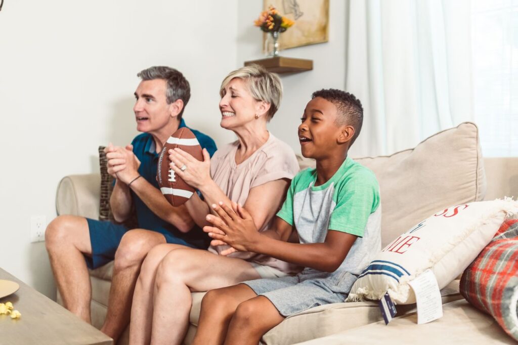 A joyful family watches a football game on the sofa, cheering with a football in hand.